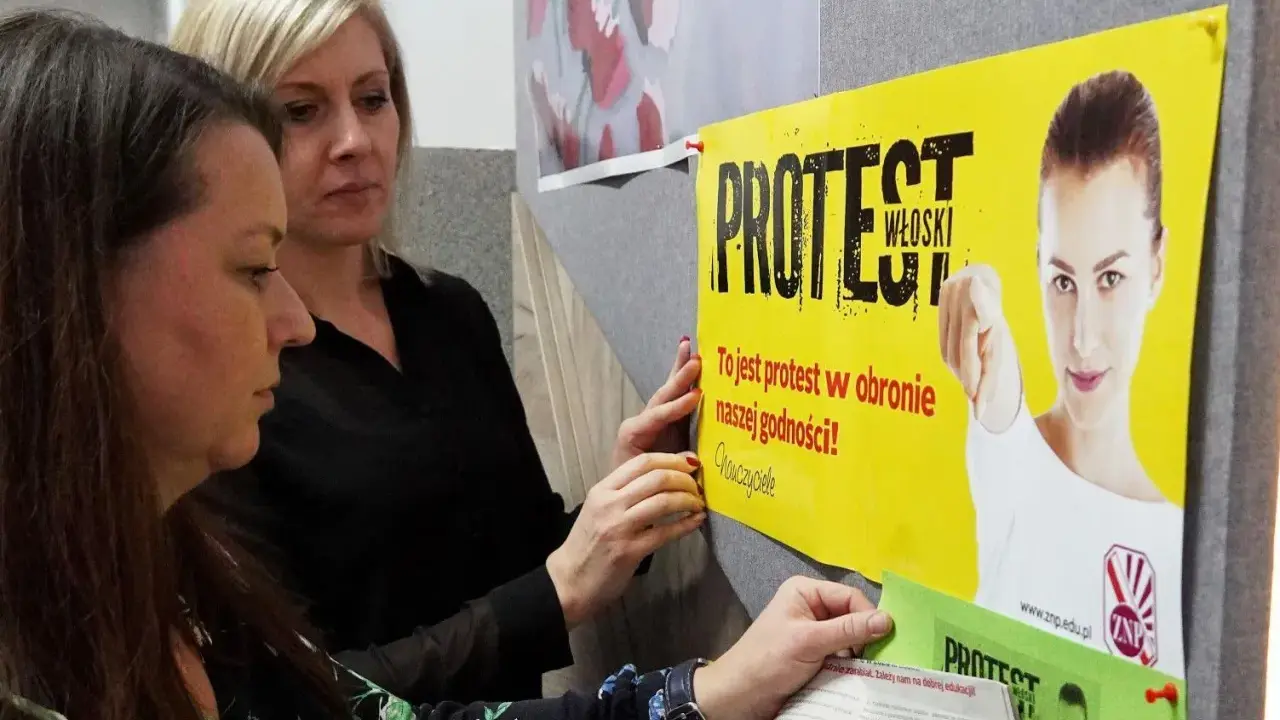 Dwie kobiety wieszają plakat "PROTEST WŁOSKI" na tablicy. Hasło głosi: "To jest protest w obronie naszej godności!".