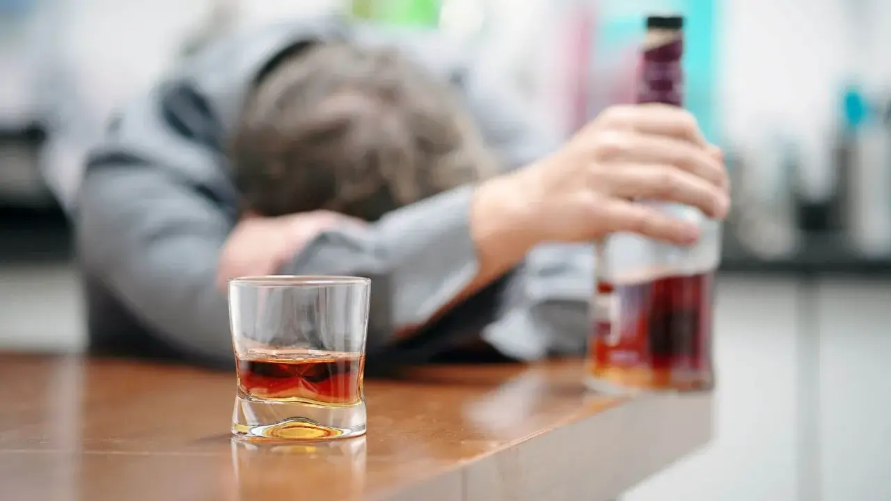 Zaniedbany mężczyzna opiera się o stół, obok szklanka z alkoholem i butelka. To obraz przymusowego leczenia alkoholika, który stracił kontrolę.
