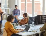 Zespół dyskutuje o regulaminie wynagradzania w biurze. Pracownicy siedzą przy biurkach, pracując na laptopach i komputerach.