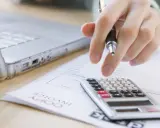 Ręka z długopisem nad kalkulatorem i laptopem. Trwają prace nad odpisem na ZFŚS 2025.