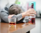 Zaniedbany mężczyzna opiera się o stół, obok szklanka z alkoholem i butelka. To obraz przymusowego leczenia alkoholika, który stracił kontrolę.
