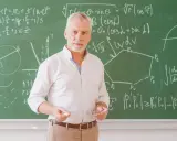 Nauczyciel matematyki z kredą i okularami przed tablicą pełną wzorów. Może to być jego urlop zdrowotny, czas na odpoczynek od nauczania.