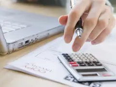 Ręka z długopisem nad kalkulatorem i laptopem. Trwają prace nad odpisem na ZFŚS 2025.