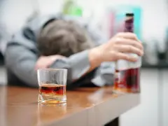 Zaniedbany mężczyzna opiera się o stół, obok szklanka z alkoholem i butelka. To obraz przymusowego leczenia alkoholika, który stracił kontrolę.