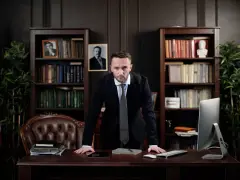 Prawnik w korporacji, stojący przy biurku, zastanawia się, ile zarabia. W tle biblioteczki i amerykańska flaga.