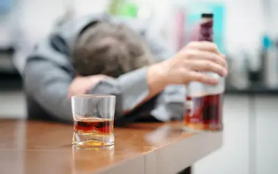 Zaniedbany mężczyzna opiera się o stół, obok szklanka z alkoholem i butelka. To obraz przymusowego leczenia alkoholika, który stracił kontrolę.