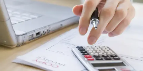 Ręka z długopisem nad kalkulatorem i laptopem. Trwają prace nad odpisem na ZFŚS 2025.