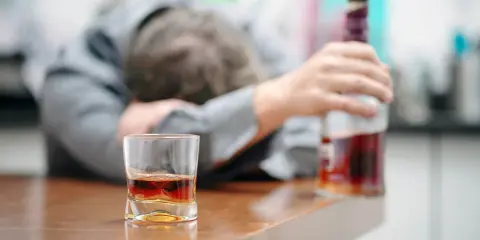 Zaniedbany mężczyzna opiera się o stół, obok szklanka z alkoholem i butelka. To obraz przymusowego leczenia alkoholika, który stracił kontrolę.