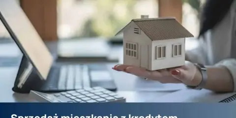 Dłoń trzyma model domu, obok kalkulator i laptop. Temat: sprzedaż mieszkania z kredytem.
