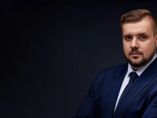 Mężczyzna w granatowym garniturze i krawacie, z zarostem, na ciemnym tle.