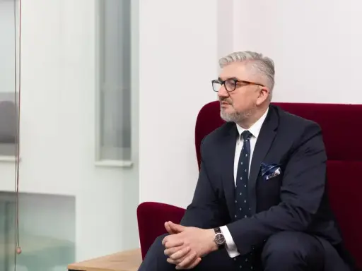 Mężczyzna w garniturze i okularach siedzi na czerwonym fotelu.