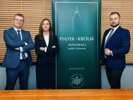 Trzech prawników w garniturach stoi przed zielonym banerem kancelarii "Piątek Królik Adwokaci".