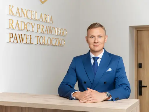 Mężczyzna w niebieskim garniturze przy biurku, za nim napis "KANCELaria Radcy Prawnego Paweł Tołoczko".