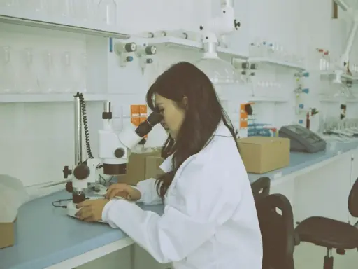Kobieta w białym fartuchu pracuje przy mikroskopie w laboratorium. Na stole leżą próbki i sprzęt.