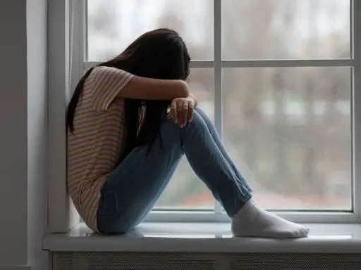Dziewczyna siedzi skulona przy oknie, ukrywając twarz w ramionach. Jej postawa sugeruje głęboki smutek, być może po traumatycznym przeżyciu, jak gwałt.
