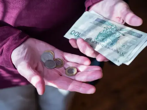 Dłonie trzymające polskie banknoty i monety. Zastanawiasz się, ile wynosi dodatek do emerytury za warunki szkodliwe?