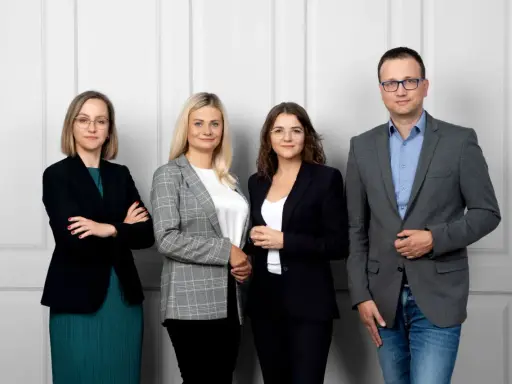 Cztery osoby w eleganckich strojach stoją przed jasną ścianą.