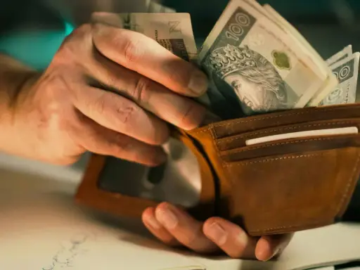 Dłonie wkładają banknoty 100 zł do brązowego portfela. To może być zapłata lub rekompensata.