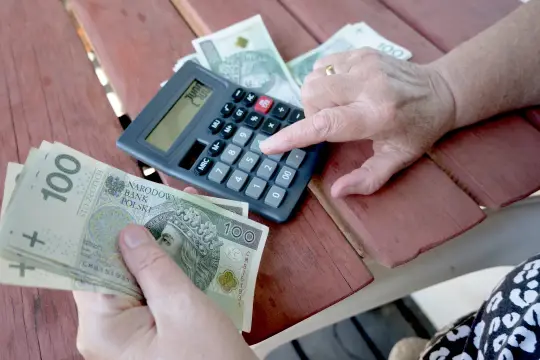 Rocznik 1952 liczy pieniądze do ZUS na wyrównanie emerytury. Kalkulator i banknoty 100 zł na stole.