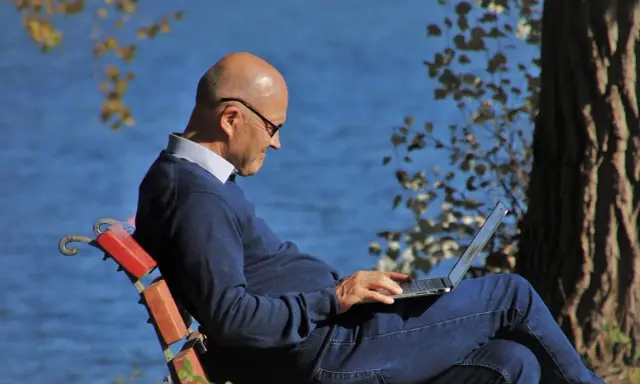Mężczyzna w okularach siedzi na ławce nad jeziorem, pracując na laptopie. Planuje swoje ubezpieczenie emerytalne, ciesząc się spokojem natury.