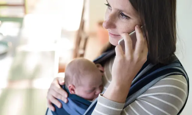 Samotna matka rozmawia przez telefon, trzymając śpiące dziecko w chuście.