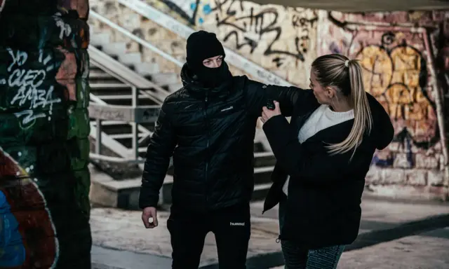 Kobieta używa gazu pieprzowego przeciwko napastnikowi w kominiarce. To przykład obrony koniecznej w niebezpiecznej scenerii z graffiti.