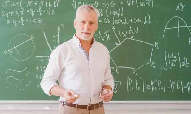 Nauczyciel matematyki z kredą i okularami przed tablicą pełną wzorów. Może to być jego urlop zdrowotny, czas na odpoczynek od nauczania.