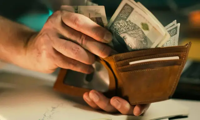 Dłonie wkładają banknoty 100 zł do brązowego portfela. To może być zapłata lub rekompensata.