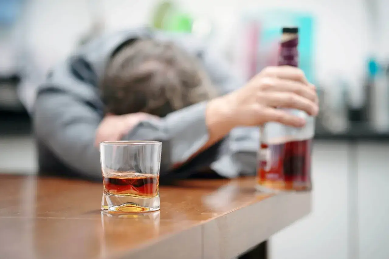 Zaniedbany mężczyzna opiera się o stół, obok szklanka z alkoholem i butelka. To obraz przymusowego leczenia alkoholika, który stracił kontrolę.