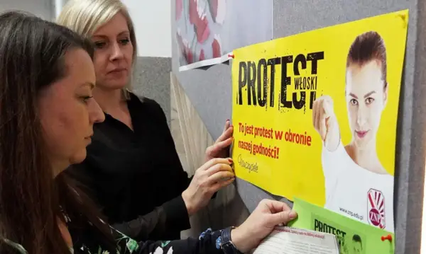 Dwie kobiety wieszają plakat "PROTEST WŁOSKI" na tablicy. Hasło głosi: "To jest protest w obronie naszej godności!".