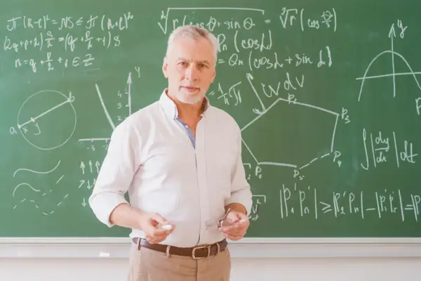 Nauczyciel matematyki z kredą i okularami przed tablicą pełną wzorów. Może to być jego urlop zdrowotny, czas na odpoczynek od nauczania.