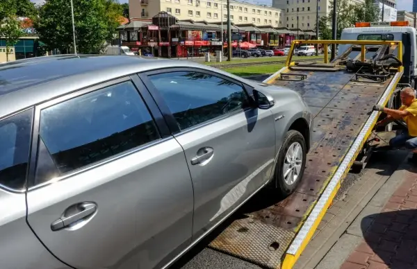 Srebrny samochód jest ładowany na lawetę. Pracownik w żółtej koszulce zabezpiecza pojazd, co może oznaczać konfiskatę samochodu.
