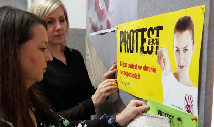 Dwie kobiety wieszają plakat "PROTEST WŁOSKI" na tablicy. Hasło głosi: "To jest protest w obronie naszej godności!".