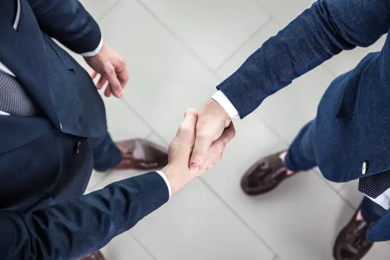 Dwaj mężczyźni w garniturach ściskają sobie dłonie na jasnej posadzce.